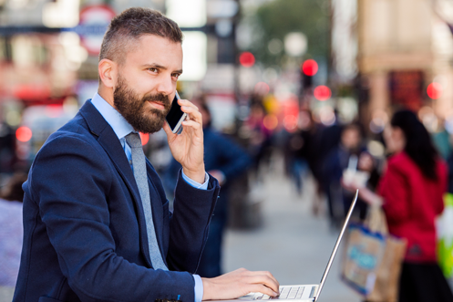 Man with beard holding a phone to his ear, in front of a laptop, in Piccadilly Circus in London, United Kingdom, wondering to himself what does the UK Economy look like for 2018
