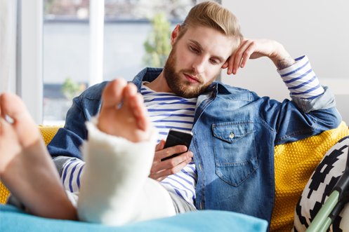 Handsome blonde man with beard, looking at phone, with broken leg, feet up, sitting on a sofa, crutch next to him, searching for Expatriate Health Insurance