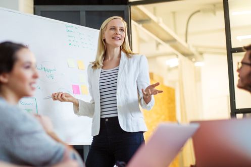 Blonde woman standing at a whiteboard, speaking to a group of people seated at a table about policy benchmarking to industry best practices
