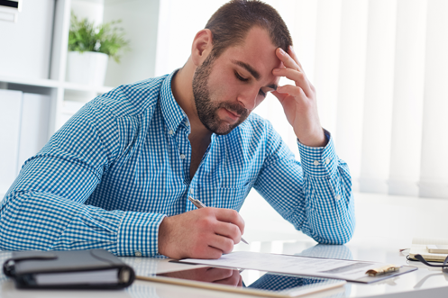 Man with beard in a blue checkered shirt, sitting at a desk, one hand on his forehead, filling out a Form I-9 with helpful nine tips
