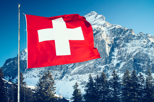 Switzerland Job Openings Switzerland Flag on flagpole in front of Swiss Alp, blue sky, indicating that Switzerland Employers Must Communicate Job Openings Occupation Types