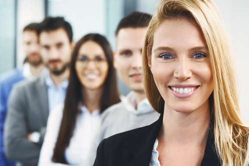 Close-up front view of mid 30's business team standing in line in their office. Focus on the closest person to the camera, smiling blond woman. There are five people total, two women and three men, as they consider Global Productivity and Employee Development