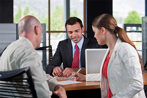 Woman in white coat and red shirt signing job acceptance papers, husband is next to her with white shirt, across from man in dark suit with red tie, take a job offer at Challenging Locations