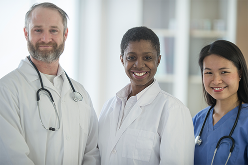 Handsome man with beard in doctor’s coat with stethoscope, two attractive women in healthcare uniforms, all three smiling as they consider relocation program models for healthcare