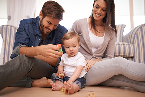Family Matters and Career Support for Spouses Impacted by Relocations smiling man with brown hair and beard wearing blue shirt, holding toy blocks, playing with child, woman with long brown hair next to him, smiling looking down at child, wearing tan sweater over white tank shirt
