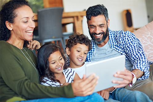 MyRelocation™ Video Resources being viewed by relocating employee and family members, one woman in green shirt, one man in blue and white checked shirt, two children, all smiling and looking at a video playing on a computer laptop