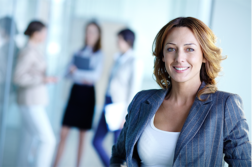 Woman with long hair, wearing blue jacket and white shirt, smiling, facing front with three other women in background, thinking about how to Recruit Top Talent