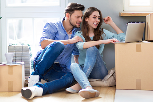 Handsome man with brown hair and beard, woman with brown hair, sitting on floor around moving boxes, looking at a computer to confirm their Traditional Temporary Housing