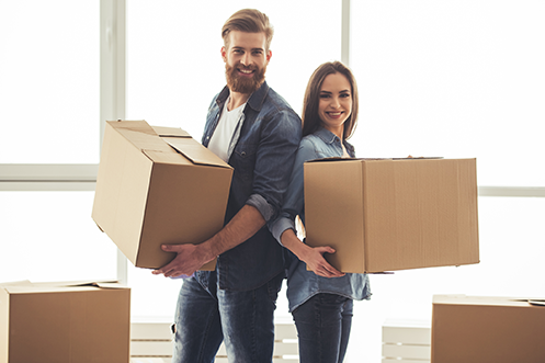 Handsome man with full beard holding a moving box, wearing a white t-shirt and blue shirt, standing next to woman with dark hair holding a moving box, happy their company chose to engage their RMC early in the relocation process