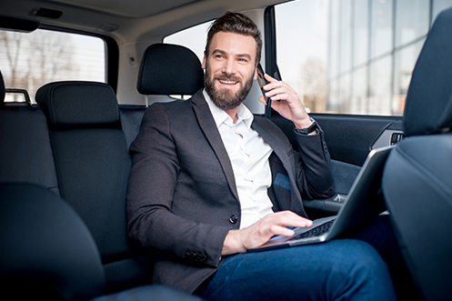 Handsome man with brown hair, mustache, and beard, riding in car, phone to ear, laptop open, smiling, talking about his company’s Expedited Bidding Process