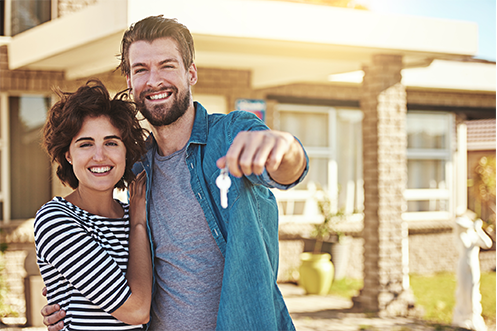Handsome man with brown hair and full beard holding keys to a house, with a big smile, wearing a gray t-shirt and blue jean shirt over the t-shirt, woman with dark hair standing to his left, able to buy house because employer gave home purchase benefits to Encourage Transferees to buy a home instead of rent