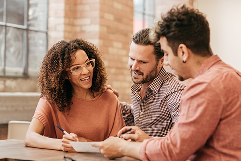 Woman with orange sweater, dark curly hair and glasses, smiling, showing new construction loan document to handsome man with dark hair and beard, wearing a checked shirt, smiling, and another man with orange shirt, curly dark hair, and beard