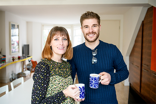 Woman with shoulder length hair and flower shirt, smiling, holding coffee mug, to left, and handsome man wearing blue sweater, smiling, with brown hair, mustache and beard, holding coffee mug, to right, thinking about their UK home and annual price growth decline