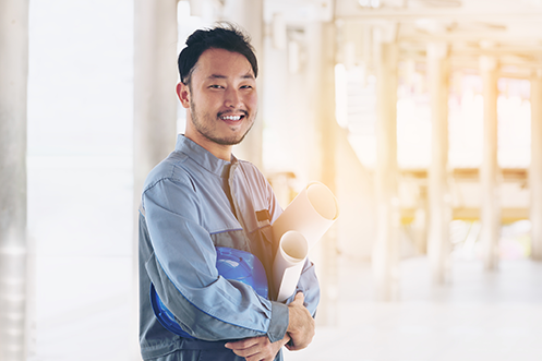 Handsome man with dark hair and beard, smiling, holding draft plans, in a factory, benefitting from a great job even during the decline of china’s labor force