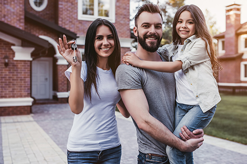 Happy family is standing with keys from their new house in hand. Smiling and looking at camera. Woman with long dark hair and white t-shirt. Handsome man with beard and gray t-shirt. Young girl with long brown hair and white shirt. The family got a new mortgage loan to buy their home