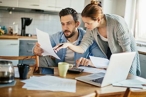 Handsome man with dark hair and beard, wearing a blue shirt and white t-shirt, holding a paper, woman leaning over and pointing at the paper, reviewing their 2017 relocation expenses