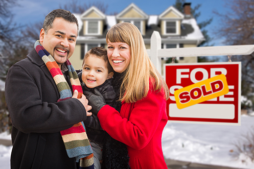 Man in coat with colorful scarf, black hair, mustache and goatee, small child, woman with blonde hair, all smiling, standing in front of house with a sold sign next to them, showing they bought this house from the home sellers