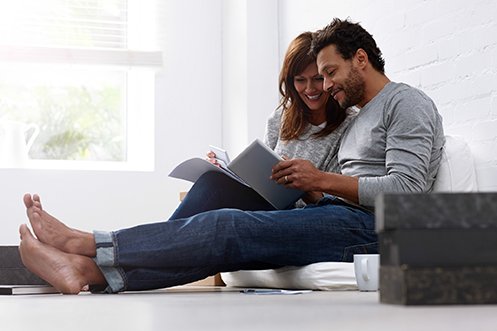 woman with long hair and man with short curly hair and beard, wearing jeans, barefoot, sitting on floor together, smiling, looking through their relocation documents prior to their home’s relocation appraisal
