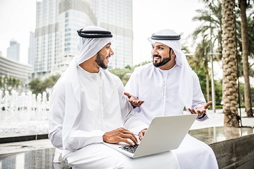 Two handsome Arabic businessmen in Dubai, smiling, talking, one holding a laptop, sitting outside in front of skyscraper, talking about how they can be successful in spite of the upcoming skills gap