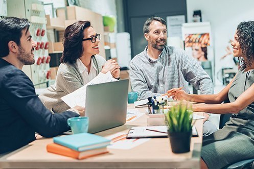 Two men and two women, all sitting at a work table facing each other, smiling, conducting market research on world economies