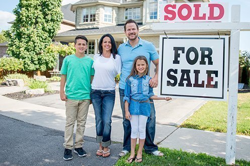Young boy in green shirt, woman in white shirt with long dark hair, handsome man with beard wearing light blue shirt and white sandals, young girl in dark blue shirt, standing next to For Sale and Sold sign, family members in front of home they purchased through a relocation company’s buyer value option program