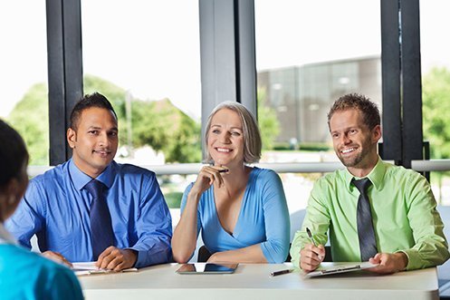 Three employees including a man in blue shirt, woman in blue shirt, and man in green shirt, interviewing a candidate in the 2019 USA job market