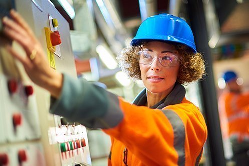 Woman in hard hat, wearing safety glasses, curly hair, reaching over to a power plant panel, happy she has one of the fastest growing Louisiana Jobs