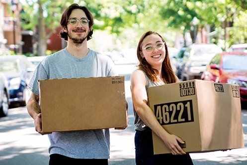 Man with dark hair, glasses, beard and mustache, smiling, carrying a box, woman with red hair, glasses, smiling carrying a box, helping out with Summertime Household Goods Moves