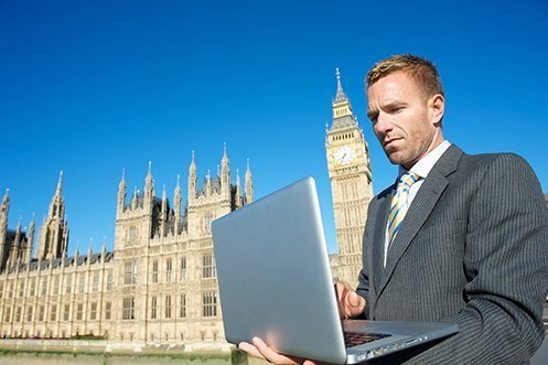 Male banker holding laptop in front of Big Ben in the background, wearing a suit, looking at laptop, planning on relocating UK workers