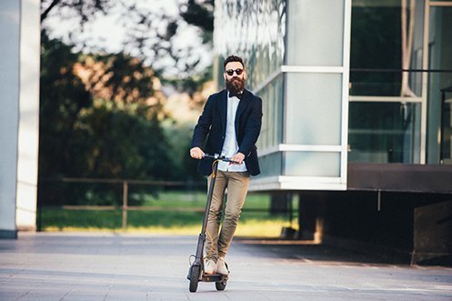Handsome man with beard, mustache, and sunglasses, wearing a bow tie, white shirt, and jacket, facing forward, riding an e-scooter in compliance with new Los Angeles city regulations
