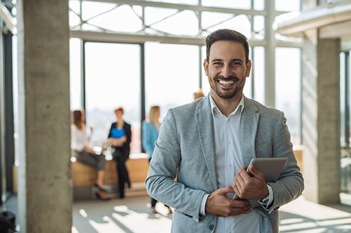Handsome man with dark hair, mustache and beard, smiling, wearing white shirt and gray jacket, holding a tablet computer, happy that he got a new job through the growth in European employment strategy initiatives