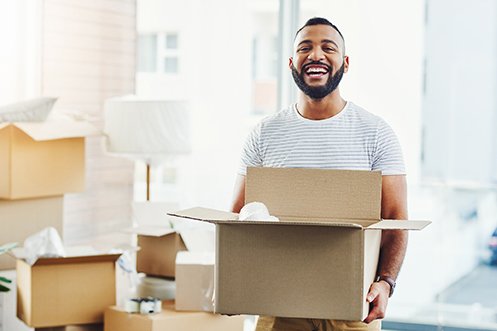 Man with dark hair, beard, wearing white striped t-shirt, holding a cardboard box, happy recipient of several Lump Sum Relocation Packages as he has progressed early in his career