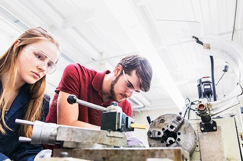 Blonde Gen Z woman wearing safety glasses, Gen Z man with dark hair and beard wearing safety glasses, both working at manufacturing industry opportunities
