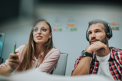 Woman with long hair and glasses, holding a pen, handsome man with dark hair and beard, wearing headphones and a wristwatch, both looking forward, working together during their domestic short term assignments
