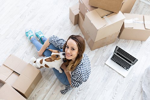 Woman with dark hair, looking up from floor with moving boxes and computer, dog on her lap licking her face, using relocation technology features to help her move