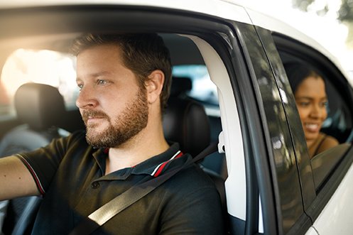 Handsome male car driver at wheel of white shared ride car, facing forward, with brown hair, beard, and mustache, wearing a black shirt, woman passenger in back seat looking out the window, smiling