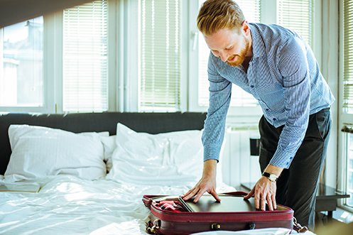 Male traveler with red hair and beard, wearing dark pants and blue shirt, packing his suitcase and taking prudent business traveler health precautions