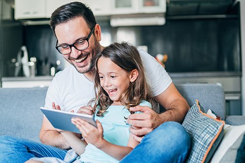 Smiling father with glasses, dark hair, and wearing a white shirt, holding young smiling daughter with long dark hair, both looking at GMS Video Destination Spotlights as they consider a relocation opportunity