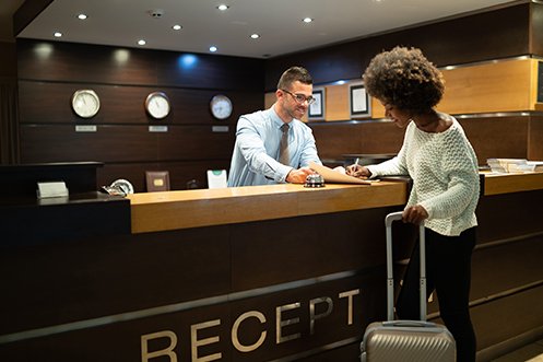Man with short hair and glasses, smiling, a front hotel desk worker helping a female business traveler with suitcase and white sweater check in to her room, the man is a new hire as noted in the January 2020 jobs report