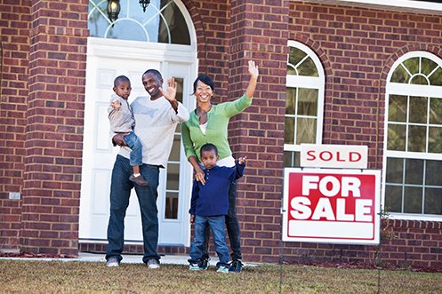 Smiling father and mother with two children, waving happily in front of their home and grateful for bvo program benefits, for sale sign with sold placard in front, brick house behind