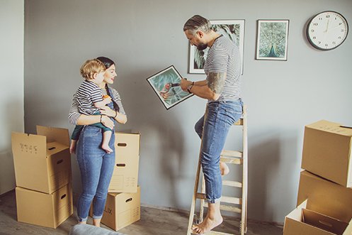 Man wearing blue jeans and striped t-shirt, with gray hair and dark beard, standing on a ladder, woman with dark hair holding a child in her arms, man is hanging photos on wall after their move during COVID COVID-19