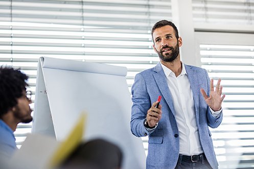 Handsome man with dark hair, mustache, and beard, wearing a white shirt and blue jacket, providing successful language training to group of students