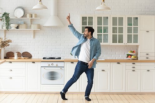 Handsome man with dark hair, mustache and bears, wearing blue jeans, white t-shirt and blue shirt, black socks, no shoes, dancing in kitchen, working from home and doing his best at beating isolation