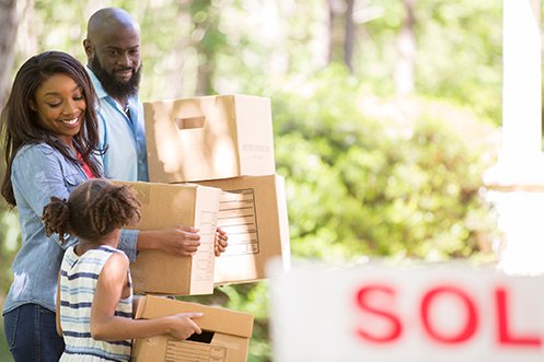 Handsome husband with beard, beautiful wife with long dark hair, both wearing blue shirts, looking at their young daughter, smiling, carrying boxes and bags during their summer move
