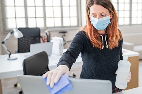 Woman with long brown hair, wearing face mask and gloves, cleaning a computer screen as she prepares for her company’s returning workforce