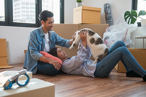 Man sitting on floor, woman laying on floor, holding up family cat, moving boxes around them, they just moved into their new apartment and had to arrange animal transportation so their cat could be with them as they flew from one state to another for a new job