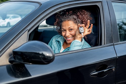 Woman driver in car with curly hair, wearing a blue shirt, holding up keys, smiling and happy with her flexible transportation solution