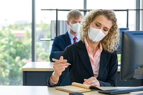 One man and one women, both working, sitting at desks, able to return safely to their workplace, wearing masks, with appropriate distance between their workspaces