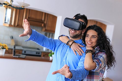 Man with dark hair, wearing blue shirt and 3D headset, woman with dark hair, smiling behind him, both looking at a home using a virtual experience