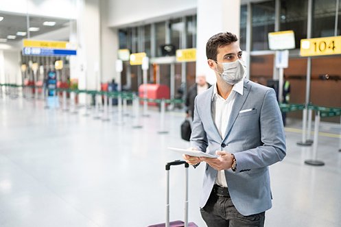 Man wearing mask at airport, with blue jacket, dark pants, white shirt, looking at a tablet, next to luggage, with dark hair, determine his best option for airline travel